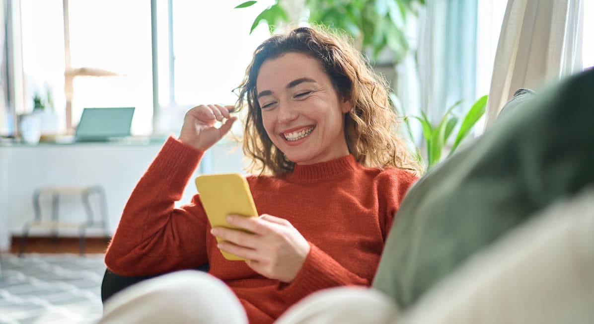 Happy relaxed young woman sitting on couch using cell phone, smiling lady laughing holding smartphone, looking at cellphone enjoying doing online ecommerce shopping in mobile apps or watching videos.